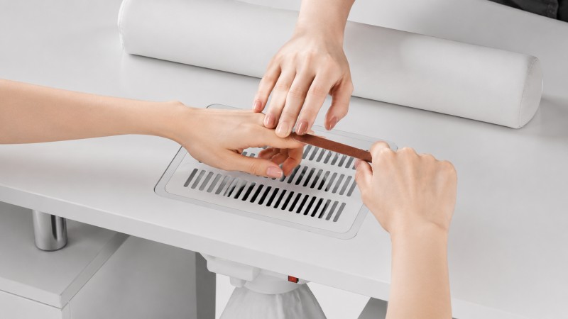 Built-In Nail Dust Collector During Manicure Filing Close-up of a nail technician filing a client’s nails directly above the ventilation grille of a built-in manicure table dust collector.